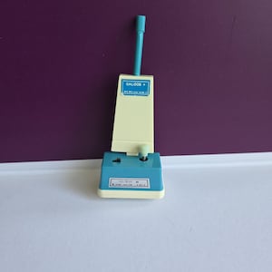 May include: A miniature toy vacuum cleaner in cream and teal plastic. The upright body features a blue label with the text GALOOB, and the base includes a smaller label with the text TRIM BEAM. It has a tall teal handle and a small teal knob.