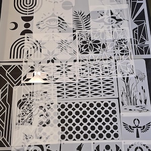 May include: A collection of translucent plastic stencils featuring various designs including geometric patterns, celestial motifs, an eye, a scarab beetle, an ankh, a seated figure, a crane in reeds, and landscape scenes, all arranged on a dark surface.