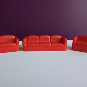 May include: A set of three miniature red plastic toy sofas, consisting of one long three-seat sofa and two smaller single-seat armchairs. Each piece features a textured surface, rounded armrests, and a solid, molded construction for play.