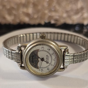 May include: A silver-tone analog wristwatch with an expansion band. The round white dial features black Arabic numerals, three hands, and a moon phase sub-dial with a dark blue sky and gold stars. The text "watch-it" and "JAPAN MOVT" are visible.