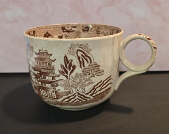 May include: A cream-colored ceramic teacup featuring a brown transferware pattern of a pagoda, trees, and landscape. The interior rim displays a decorative border, and the handle is adorned with a matching brown floral or geometric motif.