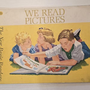Könnte beinhalten: Ein Vintage-Kinderbuch mit dem Titel We Read Pictures aus der Reihe The New Basic Readers. Das Cover zeigt eine Illustration von drei Kindern, die auf grünem Gras liegen und ein offenes Buch mit bunten Zeichnungen betrachten.