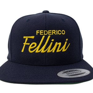 May include: A navy blue snapback hat with the name "FEDERICO Fellini" embroidered in yellow. The hat has a flat brim and a green underside.