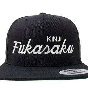 May include: Black snapback hat with the name "Kinji Fukasaku" embroidered in white script. The hat has a flat brim and a green underside. The hat is a Yupoong brand.