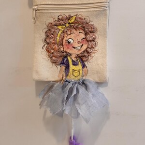 May include: A beige canvas crossbody bag featuring a printed illustration of a curly-haired character wearing a yellow headband, a dark blue shirt, and yellow overalls. A grey fabric tutu is attached to the bag, with dangling purple shoes.