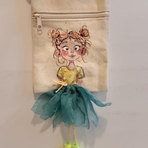 May include: A beige canvas crossbody bag featuring an embroidered illustration of a person with orange hair in buns, wearing a yellow shirt. The bag includes a teal fabric skirt overlay, yellow string legs, and small yellow boots attached at the bottom.