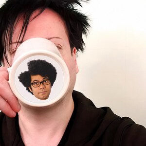 K&ouml;nnte beinhalten: Ein wei&szlig;er Keramikbecher mit einem kreisf&ouml;rmigen Grafikdruck einer Person mit dunklem, lockigem Haar und einer schwarz umrandeten Brille. Der Becher hat einen glatten, zylindrischen K&ouml;rper mit einem seitlichen Henkel f&uuml;r Getr&auml;nke.