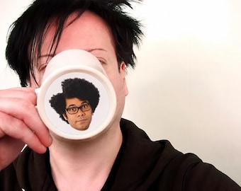 May include: A white ceramic mug featuring a circular graphic print of a person with dark curly hair and black-rimmed glasses. The mug has a smooth, cylindrical body with a handle on the side, designed for holding hot or cold beverages.