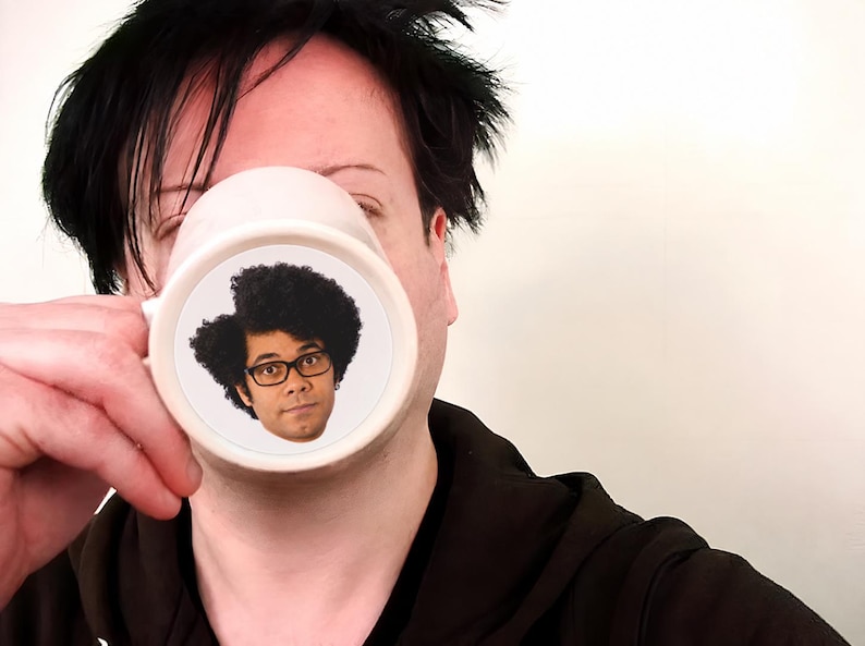 K&ouml;nnte beinhalten: Ein wei&szlig;er Keramikbecher mit einem kreisf&ouml;rmigen Grafikdruck einer Person mit dunklem, lockigem Haar und einer schwarz umrandeten Brille. Der Becher hat einen glatten, zylindrischen K&ouml;rper mit einem seitlichen Henkel f&uuml;r Getr&auml;nke.