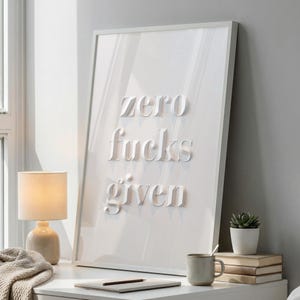 May include: A framed white art print with the words "zero fucks given" in a bold, serif font. The artwork is displayed on a white nightstand with a lamp, books, and a small plant. The overall aesthetic is minimalist and modern.