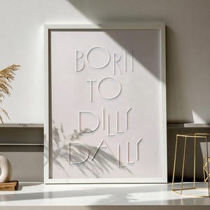 May include: White framed art print with the words "BORN TO DILLY DALLY" in a raised, minimalist design. The artwork is displayed on a white surface, with a shadow cast across the print. A gold geometric side table is in the background.