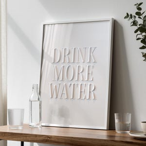 Puede incluir: Un estampado enmarcado en blanco con las palabras "DRINK MORE WATER" en letras en relieve. Una botella de vidrio transparente y dos vasos para beber se encuentran sobre una superficie de madera frente al estampado. La escena está bien iluminada y es minimalista.