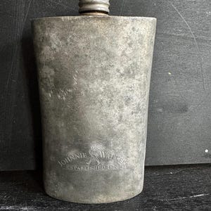 Puede incluir: Un frasco de metal vintage con tap&oacute;n de rosca. El frasco tiene forma rectangular con bordes redondeados y una superficie texturizada. Las palabras "Johnnie Walker" y "Established 1820" est&aacute;n grabadas en la parte delantera.
