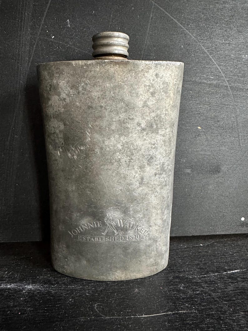 Puede incluir: Un frasco de metal vintage con tap&oacute;n de rosca. El frasco tiene forma rectangular con bordes redondeados y una superficie texturizada. Las palabras "Johnnie Walker" y "Established 1820" est&aacute;n grabadas en la parte delantera.