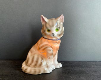 May include: A ceramic cat figurine in a seated pose. The cat features a light brown and grey striped pattern, green eyes, and a blue collar. The figurine is placed on a dark surface.