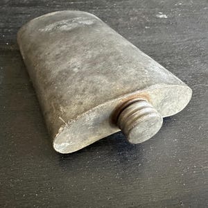 Puede incluir: Un frasco met&aacute;lico vintage, de forma ovalada, con tap&oacute;n de rosca. El frasco tiene un aspecto gris desgastado, lo que sugiere edad y uso. El tap&oacute;n es de un tono gris m&aacute;s oscuro. El frasco est&aacute; sobre una superficie oscura.