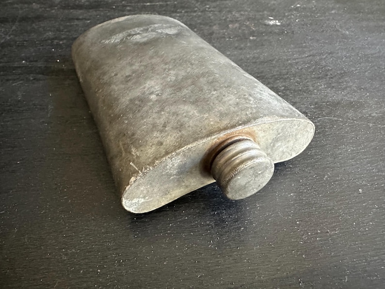 Puede incluir: Un frasco met&aacute;lico vintage, de forma ovalada, con tap&oacute;n de rosca. El frasco tiene un aspecto gris desgastado, lo que sugiere edad y uso. El tap&oacute;n es de un tono gris m&aacute;s oscuro. El frasco est&aacute; sobre una superficie oscura.