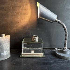Puede incluir: Una l&aacute;mpara de escritorio de metal antiguo con cuello flexible ilumina una escena con un frasco vintage, una peque&ntilde;a caja de vidrio y un recipiente plateado. Los objetos est&aacute;n dispuestos sobre una superficie oscura.