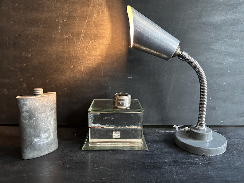 Puede incluir: Una l&aacute;mpara de escritorio de metal antiguo con cuello flexible ilumina una escena con un frasco vintage, una peque&ntilde;a caja de vidrio y un recipiente plateado. Los objetos est&aacute;n dispuestos sobre una superficie oscura.