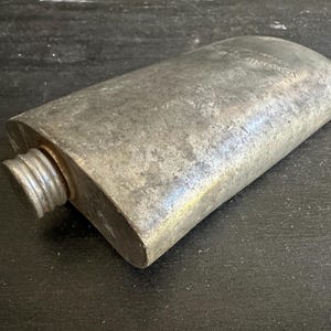 Puede incluir: Una petaca met&aacute;lica rectangular vintage con tap&oacute;n de rosca. La petaca tiene un acabado gris plateado desgastado y bordes redondeados. El tap&oacute;n es acanalado. La petaca parece ser de metal.