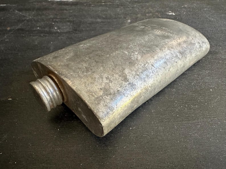 Puede incluir: Una petaca met&aacute;lica rectangular vintage con tap&oacute;n de rosca. La petaca tiene un acabado gris plateado desgastado y bordes redondeados. El tap&oacute;n es acanalado. La petaca parece ser de metal.