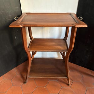 May include: A vintage wooden serving cart with two shelves and a rectangular top. The cart has curved legs and brass handles on the top. The wood has a warm, brown tone. The cart is approximately 76 cm tall.