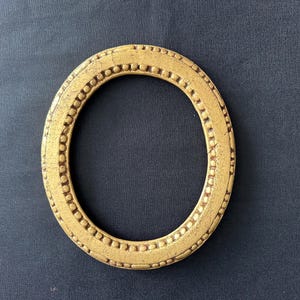 Op de afbeelding: Een ovaalvormige fotolijst met een metallic gouden afwerking. De lijst is voorzien van een decoratief randpatroon bestaande uit een herhalende reeks van kleine verhoogde stippen en korte horizontale streepjes langs de gehele rand.