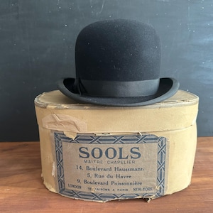 Peut inclure: Un chapeau melon noir avec un ruban assorti posé sur une boîte à chapeau ovale en carton beige. La boîte présente une étiquette avec le texte SOOLS MAITRE CHAPELIER, 14 Boulevard Haussmann, 5 Rue du Havre, 9 Boulevard Poissonnière, LONDON, PARIS, NEW-YORK.