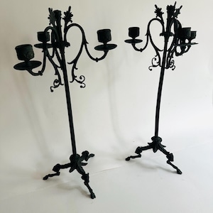May include: Two black metal candelabras with ornate, scrolling designs. Each candelabra has multiple arms to hold candles, with a decorative base and three-footed stand. The candelabras are tall and elegant.