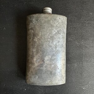 Puede incluir: Una petaca met&aacute;lica rectangular vintage con tap&oacute;n de rosca. La petaca tiene un acabado gris desgastado y el texto "JOHN WALKER ESTABLISHED 1820" est&aacute; grabado en la parte delantera.