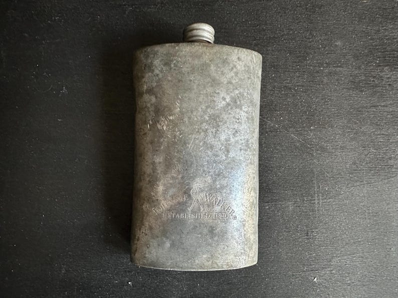 Puede incluir: Una petaca met&aacute;lica rectangular vintage con tap&oacute;n de rosca. La petaca tiene un acabado gris desgastado y el texto "JOHN WALKER ESTABLISHED 1820" est&aacute; grabado en la parte delantera.