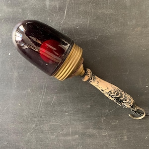 May include: Vintage dark red glass lamp with a brass base and a distressed wooden handle. The lamp has a red bulb inside and a metal hook with a black cord attached. The lamp is an antique.
