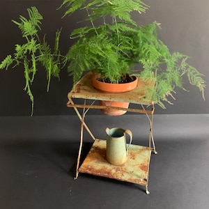 Peut inclure: Un support de plante en métal rouillé avec deux étagères, une plante en pot et un petit pichet en métal. Le support de plante est peint en vert clair et a un aspect rustique.
