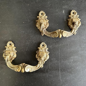 May include: Two antique brass drawer pulls with ornate, scrollwork designs. The pulls have a warm, aged patina and are set against a dark background. Each pull features a curved handle and decorative end pieces, perfect for vintage furniture restoration.
