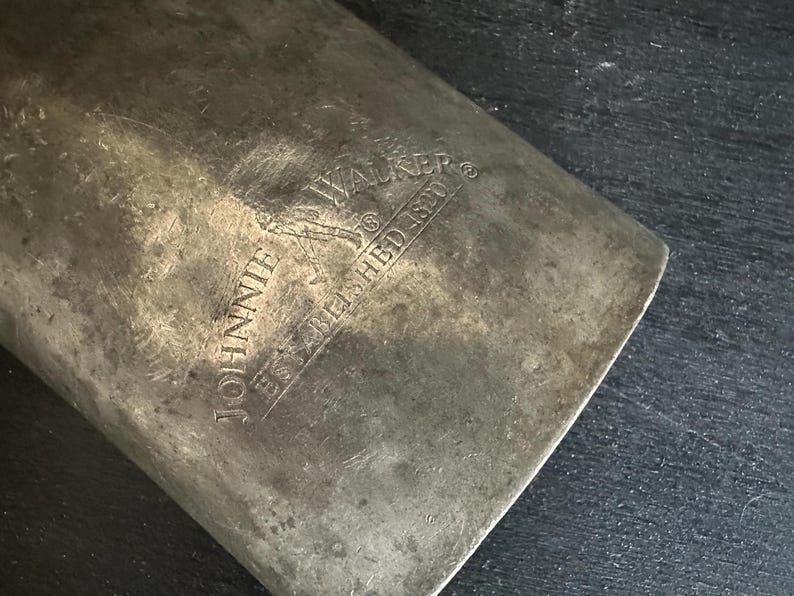 Puede incluir: Primer plano de una superficie met&aacute;lica desgastada con el texto en relieve "JOHNNIE WALKER ESTABLISHED 1820". El metal tiene un aspecto desgastado y envejecido, con ara&ntilde;azos e imperfecciones visibles.
