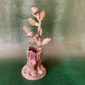 Pode incluir: Um toco de árvore de madeira decorativo com um pequeno galho de árvore crescendo do topo. O galho da árvore tem folhas e bolotas. O toco é marrom e tem uma textura áspera. A árvore é feita de metal e tem um acabamento dourado.