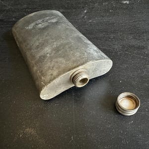 Puede incluir: Un frasco de cadera plateado con una tapa de rosca. El frasco tiene forma ovalada y una superficie texturizada, con la tapa al lado. El frasco est&aacute; vac&iacute;o y listo para ser llenado.
