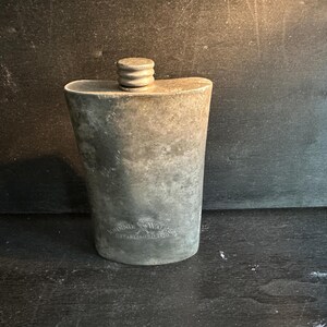 Puede incluir: Una petaca de metal envejecido con tap&oacute;n de rosca. La petaca tiene una parte superior redondeada y un cuerpo ligeramente c&oacute;nico. La superficie muestra signos de edad y desgaste, con una inscripci&oacute;n descolorida cerca de la base. La petaca es probablemente para llevar licores.