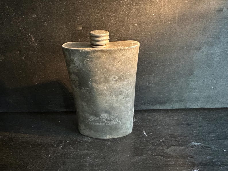 Puede incluir: Una petaca de metal envejecido con tap&oacute;n de rosca. La petaca tiene una parte superior redondeada y un cuerpo ligeramente c&oacute;nico. La superficie muestra signos de edad y desgaste, con una inscripci&oacute;n descolorida cerca de la base. La petaca es probablemente para llevar licores.