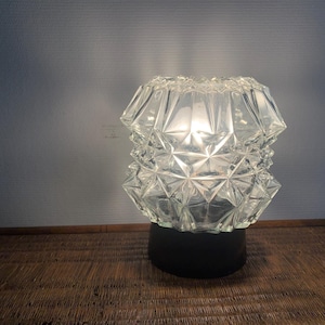 May include: A clear glass table lamp with a geometric design. The lamp has a black base and a light bulb inside.
