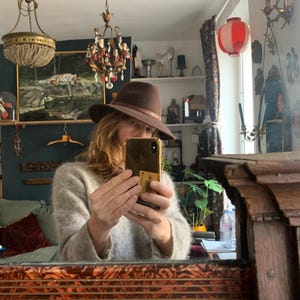 Może przedstawiać: Brązowy kapelusz typu fedora z jasnobrązową opaską. Kapelusz jest noszony przez osobę trzymającą telefon przed lustrem. Lustro odbija pokój ze starożytnym wystrojem, w tym żyrandole i dzieła sztuki.