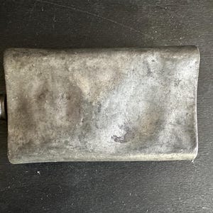 Puede incluir: Una petaca rectangular de metal desgastado con tap&oacute;n de rosca. La petaca tiene una superficie desgastada y texturizada de color gris plateado. El tap&oacute;n es de un tono gris m&aacute;s oscuro. La petaca probablemente se utiliza para transportar l&iacute;quidos.