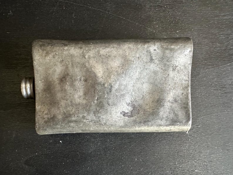 Puede incluir: Una petaca rectangular de metal desgastado con tap&oacute;n de rosca. La petaca tiene una superficie desgastada y texturizada de color gris plateado. El tap&oacute;n es de un tono gris m&aacute;s oscuro. La petaca probablemente se utiliza para transportar l&iacute;quidos.