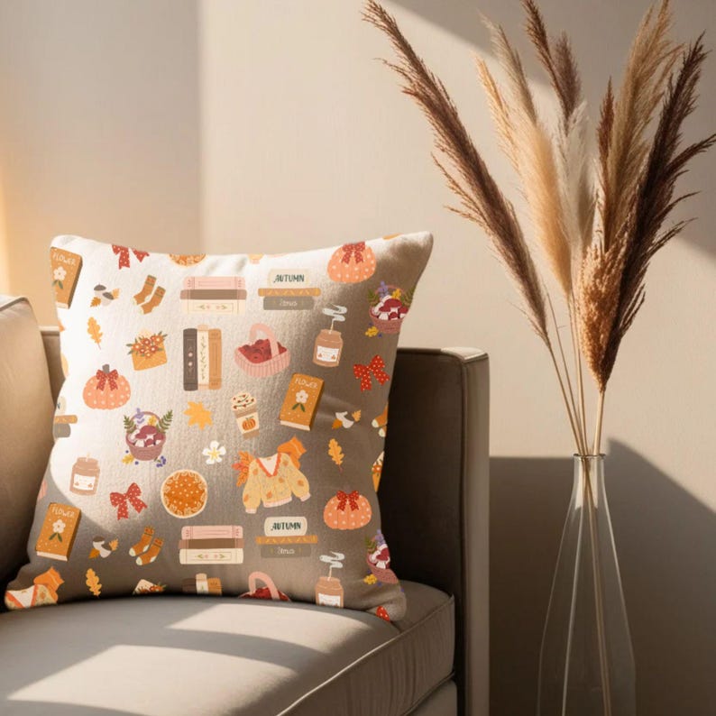 K&ouml;nnte beinhalten: Ein dekoratives Kissen mit einem herbstlichen Muster. Das Design umfasst K&uuml;rbisse, B&uuml;cher, Pullover und andere herbstliche Illustrationen in Orange-, Braun- und Beiget&ouml;nen. Das Kissen liegt auf einem grauen Sofa.