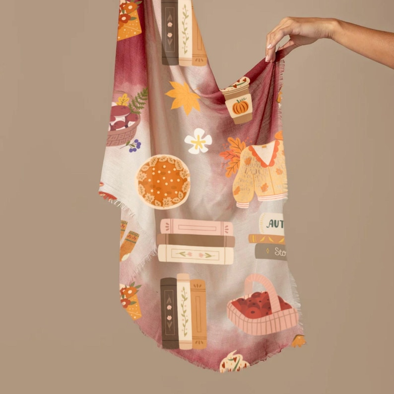 K&ouml;nnte beinhalten: Rechteckiger Schal mit herbstlichem Muster. Das Design umfasst Illustrationen von B&uuml;chern, einem Pullover, einer Kaffeetasse, einem Korb mit &Auml;pfeln und einem floralen Motiv. Der Schal hat einen Fransenrand und eine Farbpalette aus Burgunder, Creme und Orange.