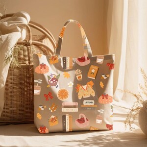K&ouml;nnte beinhalten: Beige Tragetasche mit einem herbstlichen Muster. Das Design umfasst K&uuml;rbisse, B&uuml;cher, Bl&auml;tter und andere herbstbezogene Illustrationen. Die Tasche hat eine rechteckige Form mit einem Griff.