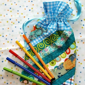 Peut inclure: Un sac à cordon en vichy bleu et blanc avec un motif patchwork. Le sac est rempli de six marqueurs colorés.