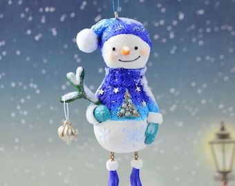 May include: A whimsical snowman Christmas ornament. The snowman wears a blue knit hat and scarf with star and tree details. It holds a sprig with a silver ornament. The snowman has a carrot nose and a smiling face. The ornament hangs from a blue string.