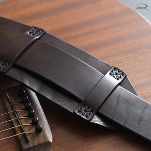 May include: A dark brown leather guitar strap with decorative stitching and embossed details. The strap is shown resting on a wooden acoustic guitar, showcasing its rich color and texture. The strap's design suggests a focus on both aesthetics and functionality.