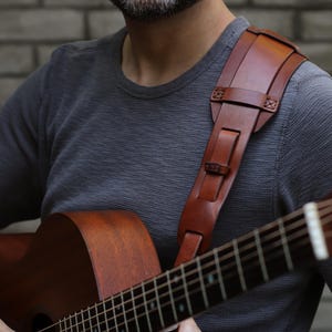 May include: A brown leather guitar strap with a shoulder pad is worn over a gray long-sleeve shirt. The strap is attached to a brown acoustic guitar. The guitar has a wooden body and a fretboard with metal strings.